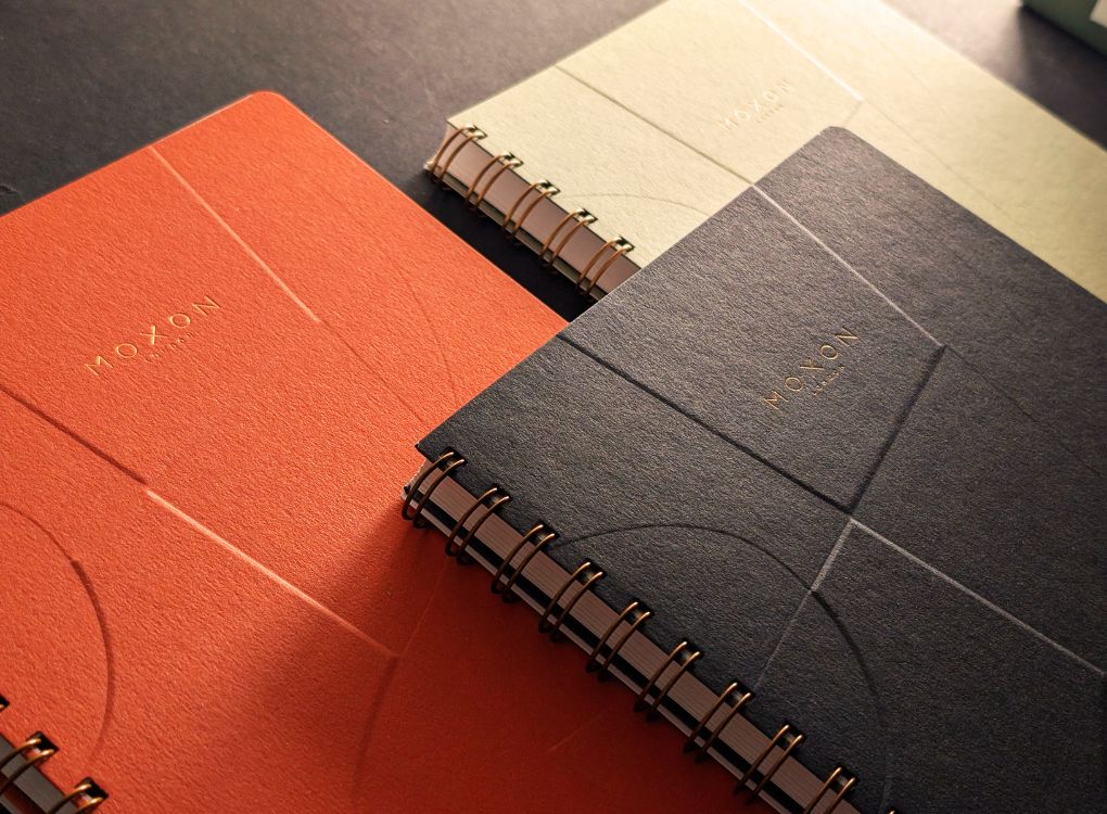 Three B5 wire-bound notebooks in Green, Orange, and Navy on a black surface. All have a blind embossed front cover.